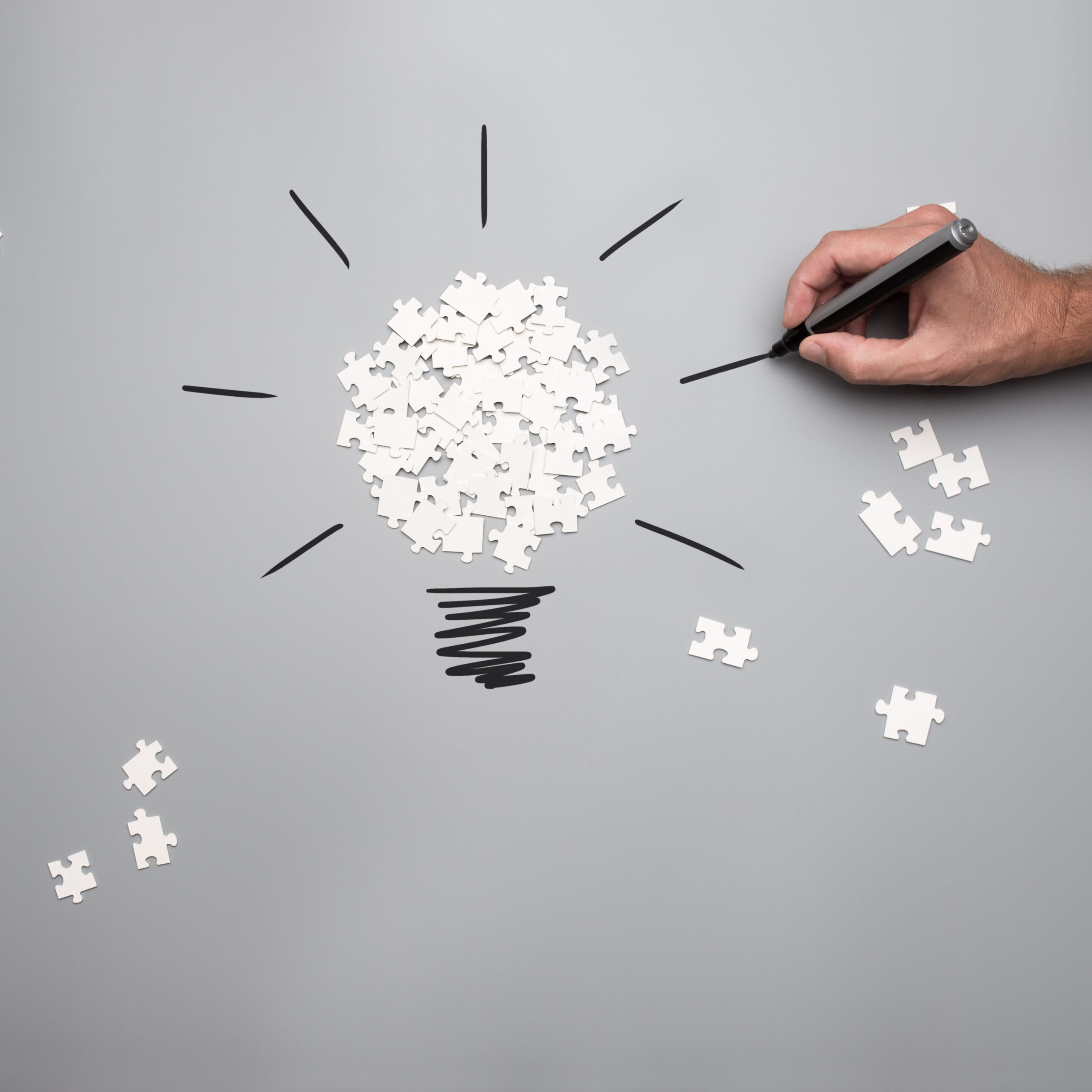 Conceptual image of business vision and idea with a pile of white scattered puzzle pieces forming a light bulb and male hand drawing rays of light on a grey background.